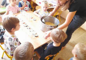 Dzieci zgromadzone wokół stołu obserwują, jak nauczycielka wkłada mus do słoika